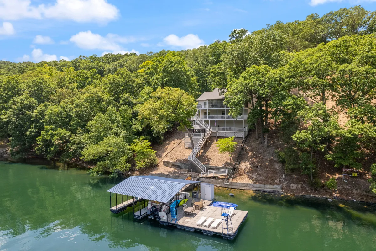 This image shows a wide exterior view of the property, featuring the house nestled on a hillside, the private dock extending into the serene lake, and lush green trees surrounding the setting. The dock is clearly visible and demonstrates how it connects directly with the house, illustrating the exclusive access to the water and the beautiful natural surroundings.