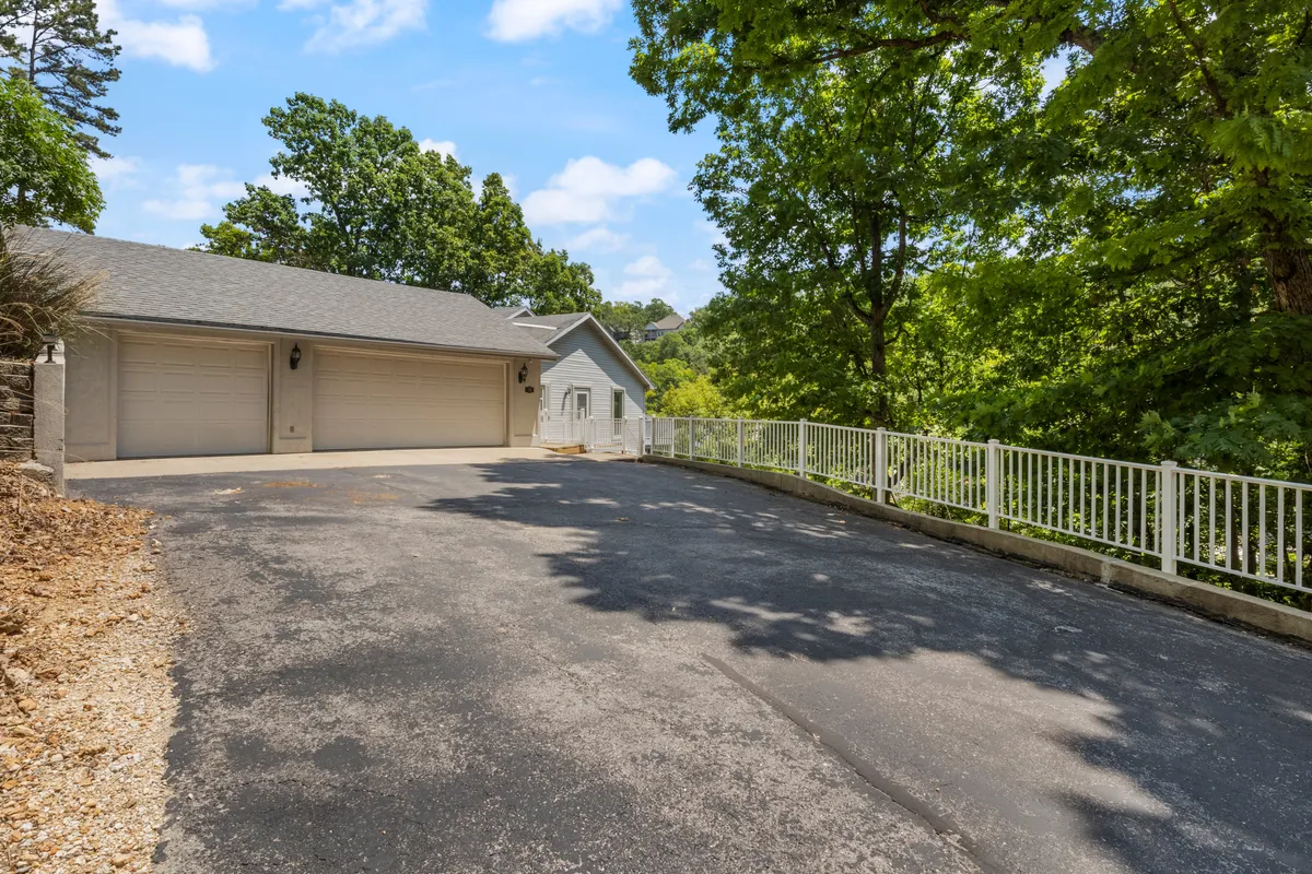 A serene exterior shot of the home's driveway, garage doors, and railings, framed by lush surrounding trees. The expansive setting hints at the welcoming entrance and outdoor space for arrivals and socializing.