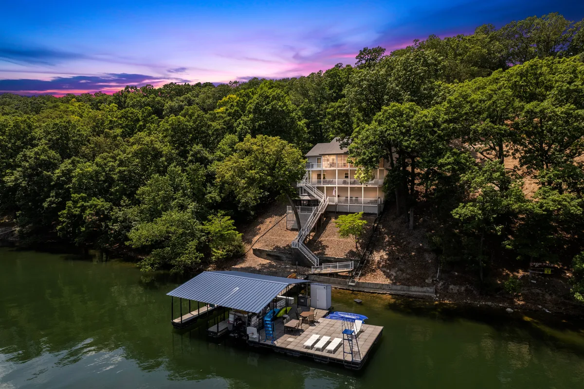 This image captures the lakeside house framed by mature trees, with a private dock leading into the gentle waters of Lake Ozark. The scene is set at sunset, bathing the dock, water, and sky in warm, golden hues. The dock is easily accessible and hints at both water activities and quiet relaxation by the lake.