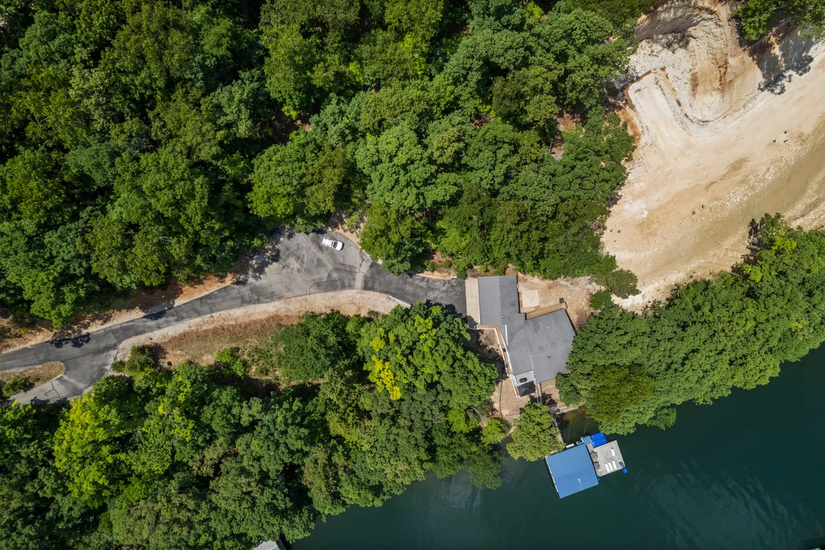 This image shows a serene lakeside setting with a house, dock, surrounding trees, and a visible road. The dock extends over the water, ideal for boating or fishing, and the overall atmosphere feels tranquil and inviting. The scene hints at both summer and fall activities with the natural landscape and accessible outdoor spaces.