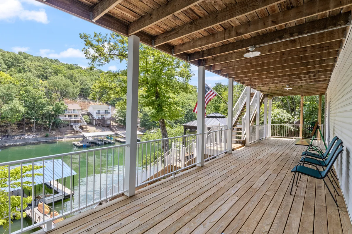 A spacious wooden deck overlooking the water, surrounded by green trees and featuring an American flag. The deck is set up for relaxing or gathering, with plenty of room to sit and enjoy the view of the lake. The water is calm and inviting, perfect for early mornings or sunset evenings.