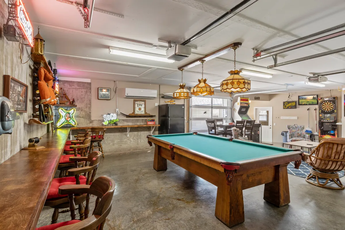 A vibrant game room featuring a pool table at its center, a bar area with stools, lounge seating, a dartboard, and colorful neon signs on the walls. The television is visible in the background, and the room is arranged to encourage both play and socializing among friends and family.