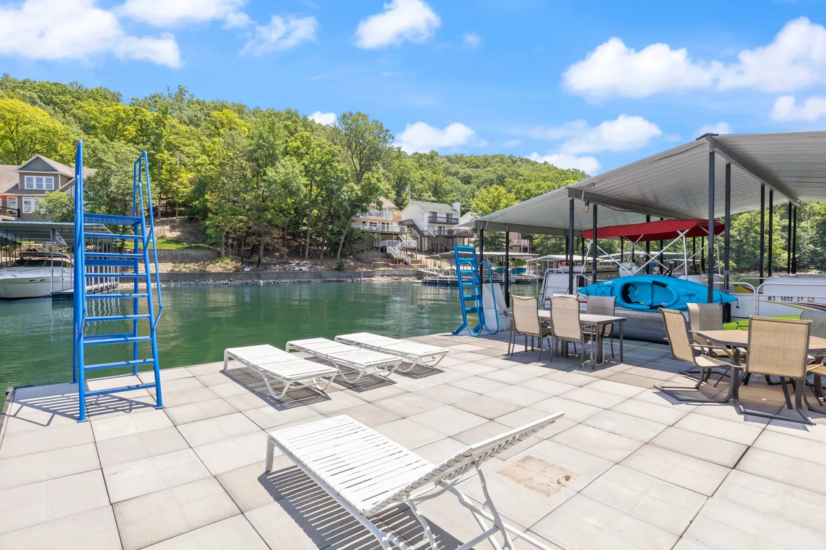 The image showcases an inviting lakefront scene with a spacious swimming dock, comfortable lounge chairs, and boats sheltered nearby. The dock extends over tranquil water, surrounded by lush trees, evoking an atmosphere perfect for outdoor leisure and water activities. The setting is bathed in warm light, hinting at stunning sunset views and relaxed gatherings by the shore.